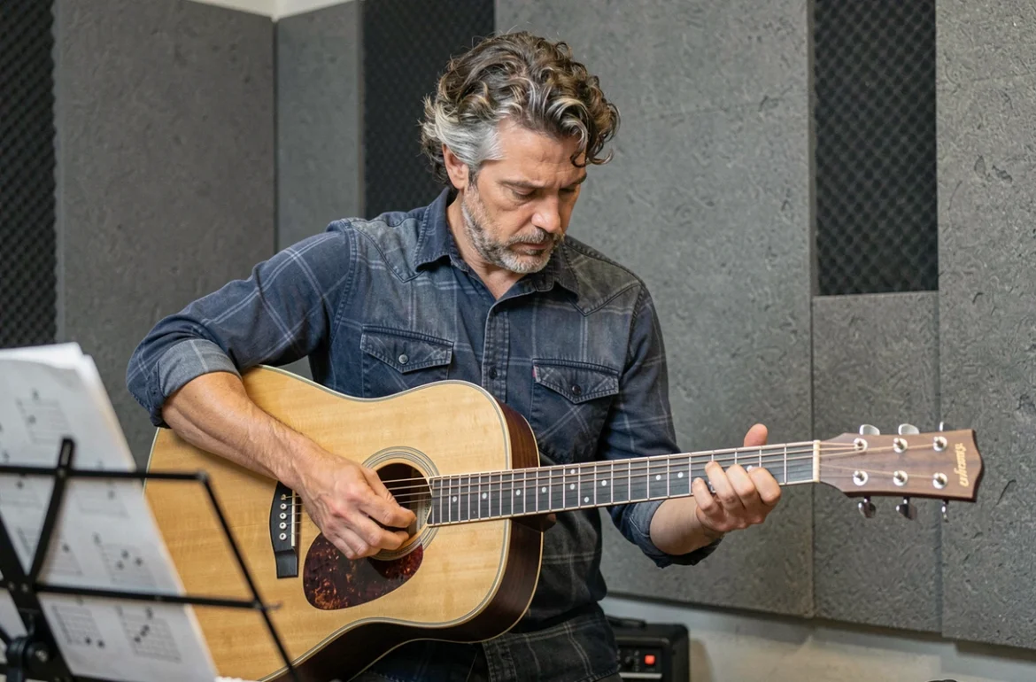 Guitariste masculin en salle de répétition analysant la position de ses doigts sur le manche d'une guitare acoustique pour perfectionner une chanson de Francis Cabrel